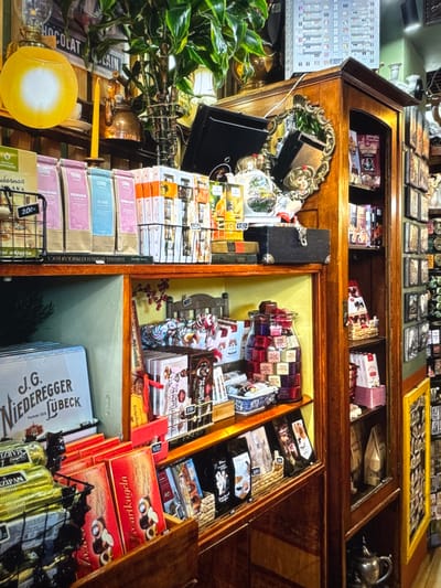 The image depicts a cluttered display of chocolates, teas, and other assorted goods within a shop. The scene is dominated by wooden shelving units and various containers filled with products. In the foreground, shelves are laden with brightly colored boxes of chocolates, bags of tea, and a large glass jar filled with individually wrapped candies. Behind these, a large light blue box with text "J.G. NIEDEREGGER LÜBECK" is visible. The shelves are packed tightly, with some items spilling over or placed precariously.
Further back, a tall wooden cabinet with glass doors showcases more items, including what appear to be packaged chocolates and possibly books or decorative objects. The upper sections of the image reveal a collection of framed pictures or prints mounted on the wall, along with hanging plants and decorative lighting fixtures. A yellow circular lamp with a diffused light source is a prominent feature in the upper left. A kettle and a single candle in a holder are also visible on an upper shelf. The overall impression is one of abundance and a somewhat chaotic, yet curated, retail environment. There are no people visible in the image. The image does not provide any information about the specific location within the city of Novinki, Russia.