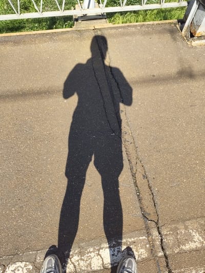 The image depicts a person's shadow cast on a paved surface, likely asphalt, in Ушаки, Russia. The shadow, elongated due to the sun's low angle, shows the silhouette of a person standing upright with arms bent at the elbows. The paved area is characterized by visible cracks and some wear. To the left of the paved surface, there is a white metal fence or railing with a green grassy area behind it. The overall lighting suggests it is daytime, with the sun casting a strong shadow. In the foreground, the feet of the person casting the shadow are visible, wearing athletic shoes.