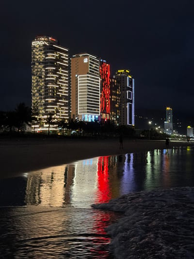 The image depicts a nighttime scene from a beach in An Hải Ward, Vietnam, showcasing a lit urban coastline. In the background, a cluster of high-rise buildings dominates the skyline, illuminated with various architectural lights. The tallest building on the left features horizontal and diagonal white/yellow light strips. Adjacent to it, a building with a light-colored facade displays horizontal white light strips and a "QBC" logo near its top. To its right, another building is brightly lit with prominent vertical red light strips. Further down the coastline to the right, additional illuminated structures are visible, including one with a yellow top. A line of dark palm trees and other vegetation forms a silhouette between the buildings and the beach.

In the midground, the wet sand of the beach acts as a reflective surface, mirroring the vibrant lights of the buildings, primarily displaying elongated white/yellow and striking red reflections. Two human figures, appearing as dark silhouettes, are walking on the wet sand. In the foreground, the ocean water gently meets the shore, reflecting the building lights in streaks of white, red, and subtle blue/purple. Small waves with white foam are visible at the very bottom right, breaking on the sand. The sky above is dark and clear.