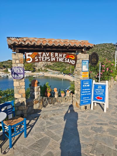 This media file captures a picturesque scene at a seaside taverna in Toroni, Greece. The foreground features a stone archway leading to a breathtaking view of a serene bay dotted with small boats. Dominating the entrance is a rustic wooden sign that reads "5 TAVERNE STEPS IN THE SAND," playfully suggesting proximity to the beach. Below this, a blue sign with white text announces "OPEN 9:00-23:00."

Further signs on the right provide more details: one highlights "TOP 100 OF GREECE 2022, 2023, 2024," indicating the taverna's recognition. Another sign, in German, translates to "WE SERVE BREAKFAST AND COFFEE WITH CAKE. HOMEMADE DISHES EVERY DAY." A prominent blue and white sign with a red arrow welcomes visitors to "THE LAST PARADISE" and directs them to "GO DOWN THE PATH."

A blue chair and a small table are visible on the left, hinting at outdoor seating. The pathway is paved with large, irregular stones, and the stone walls are adorned with decorative terracotta pots and lush green plants, some bearing pink flowers. The clear blue sky suggests a beautiful, sunny day, likely in the late afternoon or early evening, as indicated by the long shadow of a person taking the photo cast across the pathway. The overall impression is one of a charming, welcoming, and idyllic Greek holiday destination.