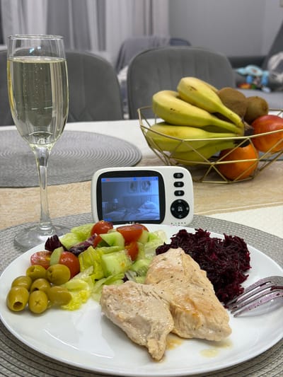 The scene captures a dining table in an indoor environment, located in Zelenaya Polyana, Russia. A white rectangular plate in the foreground contains a meal consisting of two pieces of cooked chicken breast, a portion of shredded red beetroot, a mixed green salad with lettuce, cucumber slices, and cherry tomatoes, and a cluster of green olives. A silver fork rests on the plate next to the beetroot. To the left of the plate, a clear champagne flute holds a light-colored, effervescent beverage. Centrally positioned on the table, a white baby monitor with a black screen displays a low-light image of an infant sleeping in a crib within a room, with the time "19:35" visible on the screen. The monitor features white control buttons and indicator lights on its right side. Behind the monitor, a gold-colored, geometric wire fruit basket holds a cluster of yellow bananas, two brown kiwis, and three orange-colored fruits. The table is covered with a light-colored tablecloth, and a round, woven gray placemat is visible under the champagne flute. The background is blurred, showing grey upholstered dining chairs and light-colored curtains.
