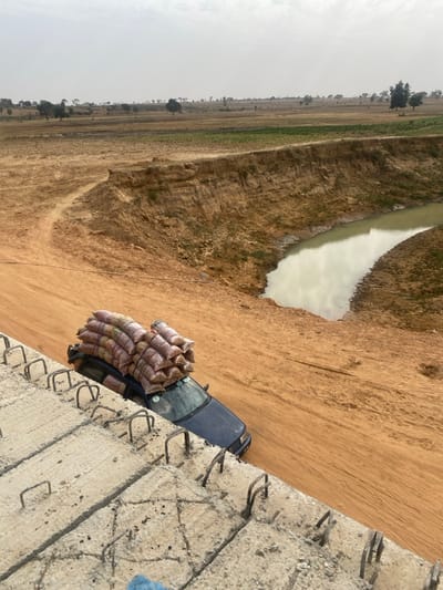 This image depicts a rural or semi-rural scene in Gyellesu, Nigeria, from an elevated perspective. In the immediate foreground, to the lower left, a rough concrete structure with exposed and bent steel rebar indicates ongoing construction. Below and to the right of this structure, a dark blue passenger vehicle, possibly a sedan, is positioned on an unpaved, reddish-brown dirt road. The vehicle's roof is heavily laden with approximately 10-12 light-colored sacks, secured in place. The dirt road curves left into the midground. To the right of the road, a significant gully or eroded bank descends to a winding body of murky, light greenish-brown water that reflects the overcast sky. The background reveals an expansive, dry landscape dominated by reddish-brown earth, sparse green vegetation, and scattered trees under a hazy, light grey sky. No individuals are visible.