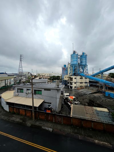 This is a medium-angle, eye-level shot of an industrial area under a cloudy sky, likely in Kaohsiung, Taiwan, given the presence of the characteristic cement silos and industrial buildings. The scene features several large, blue cylindrical silos, part of a cement or concrete plant, dominating the center-right of the frame. These silos are supported by a steel structure and connected by large, blue pipes. Adjacent to the silos is a cream-colored building with multiple windows.

In the foreground, a white cement mixer truck, with a colorful, painted drum, is parked on a dirt lot filled with aggregate. To the left of the silos, a smaller, grey building with a beige metal roof is visible. This building has multiple windows with green frames and air conditioning units. A brick wall with concrete supports runs along the front of this building and the adjacent lot.

Further in the background, a large, lattice-style electricity transmission tower stands prominently on the left side of the image, with power lines extending across the frame. Other industrial buildings, some with corrugated metal roofs and white walls, are scattered throughout the mid-ground and background, suggesting a developed industrial zone. The ground in the industrial area appears wet, possibly from rain, and a dark asphalt road with double yellow lines is visible in the lower portion of the image. The overall atmosphere is overcast and somewhat industrial. No people are clearly visible in the frame.