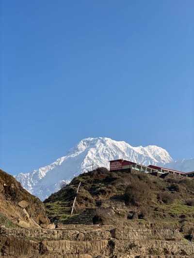 The photograph captures a scene in Lwang Ghalel, Nepal, featuring a tall snow-capped mountain juxtaposed with a cliffside structure. The sky is clear and blue. The structure is identified by a sign as "HOTEL HIMALAYAN MAJESTY." The mountain is covered in snow. The cliffside is rocky and covered in vegetation.