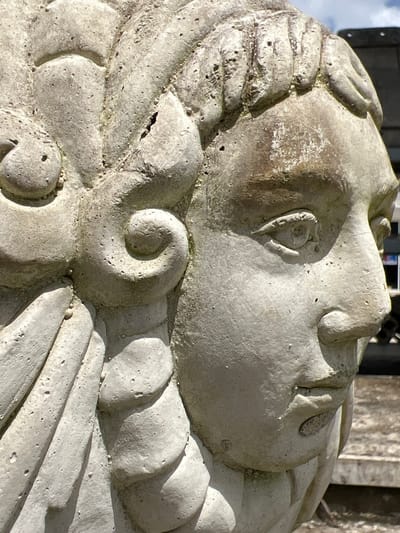 A close-up, side-profile photograph captures a weathered, light-colored concrete statue of a person's head. The statue's face is detailed with a prominent eye, nose, and mouth, showing signs of age and erosion. The hair is sculpted into curls and what appears to be a headdress or elaborate hairstyle, with some sections of the statue exhibiting cracks and pitting. The background is blurred, suggesting an outdoor setting with some hints of greenery and possibly a building structure. The lighting indicates a daytime scene with sunlight casting shadows on the statue. The statue is located in Pa Khlok, Thailand.