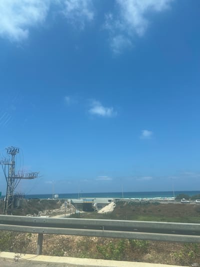 The image depicts a view of the Mediterranean Sea from a highway overpass in Israel. The sky is clear and blue with scattered white clouds. In the foreground, a metal guardrail runs along the edge of the highway. Beyond the guardrail, there is a landscape with dry vegetation and some greenery. A large electrical pylon with wires and transformers is visible on the left side of the image. In the midground, there is a construction site with an overpass bridge being built. Further back, the blue Mediterranean Sea stretches to the horizon, with a beach lined with umbrellas and people visible on the right side of the scene. Cars are seen on the highway and near the beach. The overall scene suggests a coastal area in Israel on a sunny day.