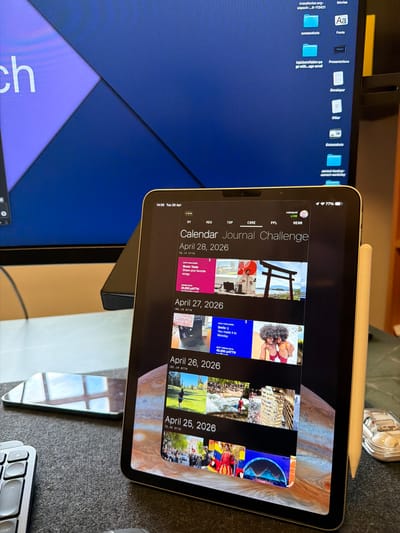 The image captures a workspace in Kafr Yasif, Israel, featuring a desktop monitor and a vertically oriented iPad on a stand. An Apple Pencil rests on the top edge of the iPad, and part of a silver keyboard is visible on the gray, textured desk surface.

The desktop monitor on the left displays a dark blue background with several file folders, including "Movies," "Fonts," "Presentations," "Developer," "Other," and "Screenshots." Other visible text on the monitor includes "transfonter.org-unpack-...8-112421," "topUsersTable+page with...age scroll," and "correct-backup-correct-workintig." Both the monitor and the iPad display "14:56 Tue 28 Apr," indicating the time as 2:56 PM on Tuesday, April 28.

The iPad's screen shows an application titled "Calendar Journal Challenge." The app interface includes tabs like "NEW," "TOP," "CORE," "PPL," "NEAR," and "LIVE," and the battery status shows 77%. The main content area displays daily challenges and user-generated content from April 2026.

For "April 28, 2026," a "DAILY CHALLENGE: Music Taste" prompts users to "Share your favorite songs," with a "WINNER GETS: 30,000 PATTN." Another challenge for this date is "Smile :) You made it to Monday." The associated images include two men with curly hair and a traditional Japanese Torii gate. "April 27, 2026" features "THANKS COLLECTIVE MEMORY" and an image of someone at a desk. "April 26, 2026" shows a person on a golf course, and "April 25, 2026" depicts a crowded street scene. Each date entry also shows engagement metrics, such as "48.1M ATTN," "102.1M ATTH," "101.IN ATTN," and "76.4M ATTN." The iPad's wallpaper is a striking image of Jupiter.