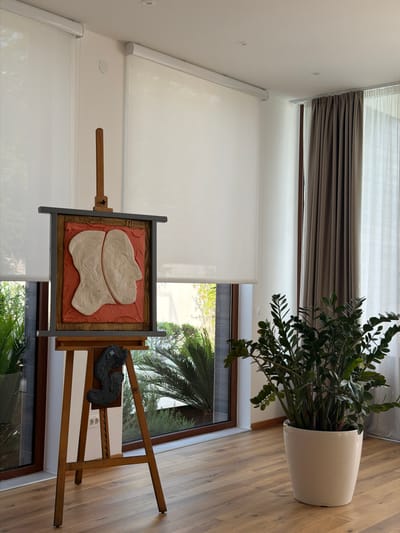 The image depicts an indoor scene in a modern dwelling in Krimovice, Montenegro. The room features large windows with white roller blinds and wooden flooring. Prominently displayed is a wooden easel holding a framed artwork. The artwork itself is a relief sculpture with two abstract, light-colored forms mounted on a textured reddish background, enclosed within a dark wooden frame. To the right of the artwork, a pair of dark, possibly leather, shoes are placed on a shelf attached to the easel. In the background, a large potted plant with lush green foliage adds a touch of nature to the interior. The lighting suggests it is daytime, with natural light filtering through the windows, highlighting the textures and forms within the scene. The overall ambiance is one of quiet contemplation, with the artwork and sculptural shoes suggesting a personal and artistic space. The scene is devoid of people, focusing solely on the arrangement of objects within the room.