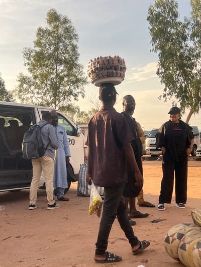 The scene depicts an outdoor gathering point in Gbaza, Nigeria, characterized by a reddish-brown dirt ground and several individuals alongside white passenger vans. The lighting suggests either early morning or late afternoon with a soft, warm hue.

In the foreground, a man walks from right to left, wearing a dark short-sleeved shirt, dark trousers, and dark sandals with white accents (one sandal displays "GUCCI"). He carries a large, circular tray on his head, laden with numerous conical, light-brown food items. His left hand holds a clear plastic bag containing yellow objects.

To the left, a white passenger van is parked with its side door open, revealing interior seating. The number "20" is prominently displayed on its side. Standing next to the van are two men: one in a light blue traditional garment and a dark cap, holding a dark bag, and another wearing a light blue long-sleeved shirt, khaki trousers, a dark grey backpack, and dark sneakers with white soles. They face each other, seemingly engaged in conversation.

Further to the right and in the mid-ground, another man wears a gold-colored long-sleeved shirt and appears to be smiling, looking towards the right. Adjacent to him, a woman stands facing left, wearing a black t-shirt, a black long-sleeved jacket, black pants, and a black baseball cap. Her dark sandals have white soles and straps, with "OUOFL" visible on one.

In the background, multiple trees with slender trunks and dense, light green foliage are visible against a clear sky with scattered white clouds. Several large, cylindrical objects with a distinct brown, tan, and black geometric pattern lie on the ground in the lower right corner. A white and blue striped woven bag rests on the ground near the man in the blue garment. A second white van is partially visible in the far right background.