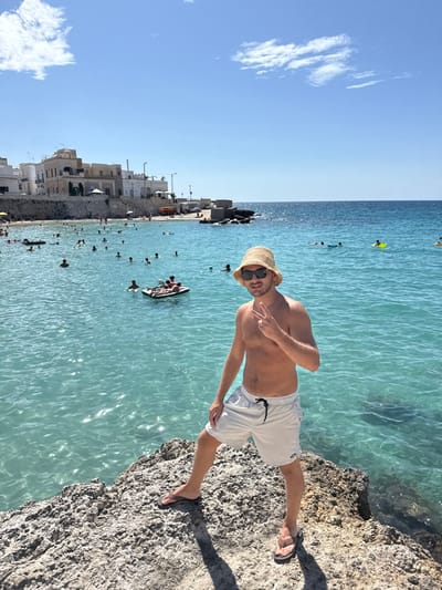 The image displays a male subject standing on a light-colored rocky outcrop in a coastal area of Nardò, Italy, under clear blue skies with scattered white clouds. The subject, shirtless, wears light-colored swim shorts, dark flip-flops, a woven straw bucket hat, and sunglasses, holding his left hand in a two-finger "V" gesture towards the camera.

The background features a bay with clear, light blue-green water. Numerous individuals, approximately 20 to 30, are visible swimming or floating in the water; one person is on a yellow inflatable ring, and two people are on a black and white inflatable raft. Along the shoreline, a collection of white and light-colored buildings, characteristic of Mediterranean architecture, are situated above a stone retaining wall and a narrow beach. A small pier or rocky extension protrudes into the water on the right side of the bay.