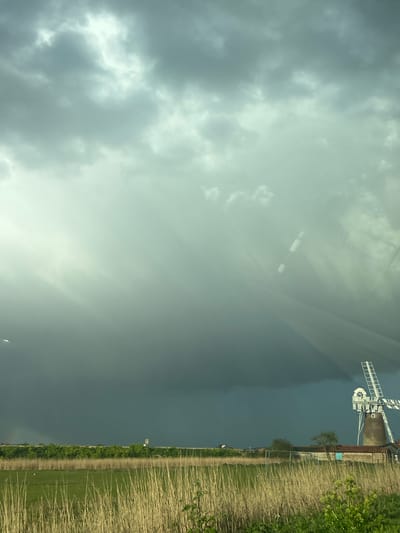 The image depicts a landscape in Broadland, United Kingdom, dominated by a grey, overcast sky and a windmill. The lower right of the frame is occupied by a field with a dirt path running along its edge, bordering a dense line of green vegetation and tall reeds. The windmill stands in the distance, its blades visible against the backdrop of the dark sky. The perspective suggests the photo was taken from a low vantage point, possibly from inside a vehicle looking out, as there are hints of reflections on the sky. The overall scene is rural and muted in color, conveying a sense of a potentially stormy or cloudy day.