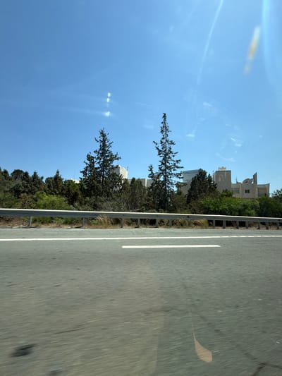 This media file captures a view from a vehicle traveling along a road in Parekklisia Community, Cyprus. The scene is dominated by a clear blue sky and a section of asphalt road with white lane markings. To the left of the road, there is a metallic guardrail separating it from a lush green area filled with trees and vegetation. In the background, several multi-story buildings are visible, peeking out from behind the trees. One of the buildings has a large blue and white sign with the text "TRILOGY" prominently displayed, along with other smaller text that is not clearly legible. The sun appears to be high in the sky, suggesting it is daytime, and the weather seems clear and sunny, with some lens flare visible in the upper left portion of the image. The overall impression is one of a typical day of travel in a suburban or semi-rural area.