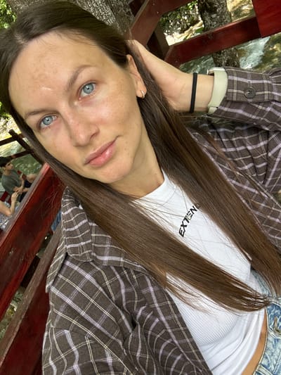 The image shows a woman with light skin and blue eyes, wearing a plaid shirt over a white ribbed tank top and denim shorts. She has long brown hair and is wearing a gold necklace that says "EXTEND" and a watch on her left wrist. She is sitting on a wooden bench next to a body of water with a rocky riverbed. In the background, two men are sitting on the same bench, with one man looking towards the camera. The environment appears to be outdoors, possibly a park or a natural scenic area. The lighting suggests it is daytime. The location of Bare Kraljske, Montenegro is not explicitly indicated in the image itself, but the description can be provided if this context is assumed.