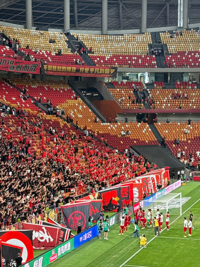 A soccer match in Longxing, China is underway. Stadium seating is colored yellow, orange, and red. Players in green, red, and yellow uniforms are on the field. Spectators wave banners displaying Chinese script. A banner reads, "No Fear."