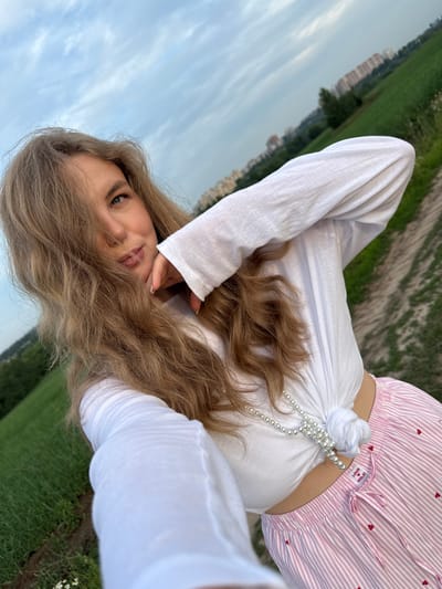 A young woman with long, wavy blonde hair is taking a selfie outdoors. She is wearing a white long-sleeved shirt tied at the waist and pink and white striped pants with small red hearts. A pearl necklace is draped around her neck and over the knotted shirt. Her left arm is raised, with her hand near her face, and her right hand is holding the phone to take the picture. The background consists of a field of green grass, a dirt path, and a cloudy sky with some buildings in the distance. The location is identified as the city of Сновицы, Russia.