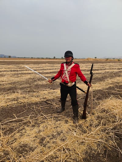 An adult male stands centrally in a harvested field in Kottalapalli, India, under an overcast sky. He wears a red military-style tunic with white cross belts and trim, dark trousers, and dark boots. A black shako-style helmet is on his head. His left hand holds a silver-bladed saber with a gold hilt, extended outward. His right hand holds a dark-brown long firearm, likely a musket or rifle, with an attached bayonet, resting on the ground beside him. A light-colored strap is affixed to the firearm. His facial expression shows a determined or strained look, with his mouth slightly downturned. The ground is dark, cracked earth covered extensively with dry, golden-brown straw stubble and stalks. The field extends into the distant background, where sparse vegetation, small trees, and low, hazy mountains are visible under a uniformly grey sky.