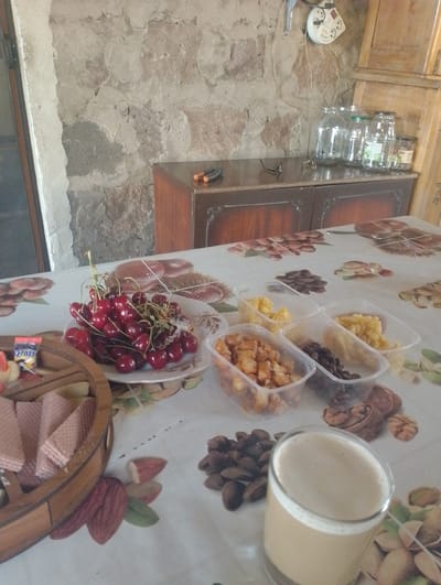 The image depicts a rustic interior scene. The background features a stone wall and a wooden cabinet with glass jars on top. In the foreground, a table is set with a variety of food items. A plate of fresh cherries is prominent, alongside several plastic containers filled with dried fruits and nuts, a wooden tray with wafer-like snacks, and a glass of foamy beverage. The overall impression is one of a casual meal or snack setting. The location in the city of Nor Geghi, Armenia, suggests a rural or semi-rural setting. The absence of people indicates a focus on the still life arrangement of the food and the environment.