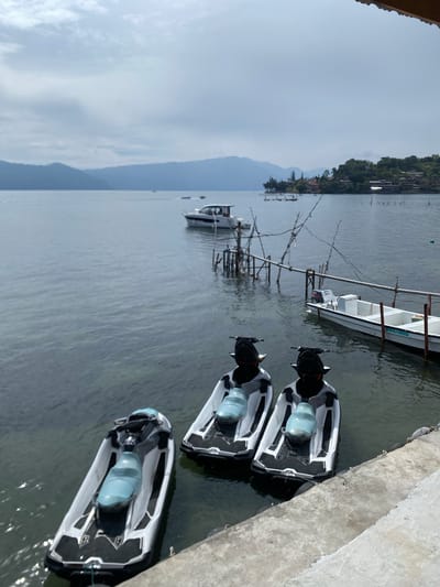 Three jet skis are docked at the edge of a body of water, with a concrete walkway in the foreground. The jet skis are white and gray with light blue seats. In the background, a small white boat is moored to a pier made of wooden poles. The water is calm, and a distant shoreline with trees and buildings is visible. The sky is overcast.