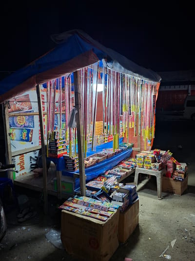 Here is a description of the image:
A roadside stall in Keudegeudong, Indonesia, is selling fireworks. The stall is constructed with wooden supports and covered with blue and orange tarps. Long sparklers are displayed on shelves, along with boxes of smaller fireworks. The stall also has boxes of drinks. The ground around the stall is paved and littered with trash. A motorbike is parked in front of the stall.