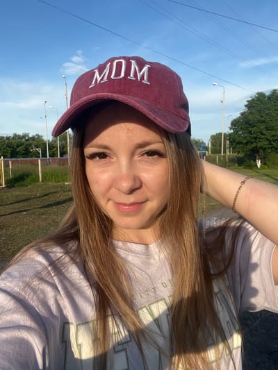 A young woman with long, blonde hair and brown eyes is taking a selfie outdoors. She is wearing a maroon baseball cap with "MOM" embroidered in white letters on the front and a light-colored t-shirt. She is touching her hair with her right hand, which has a delicate gold bracelet on her wrist. There are some pink marks on her forearm. The background suggests a semi-urban or park-like setting with a clear blue sky, streetlights, trees, and some fencing. The lighting indicates it's either early morning or late afternoon. The image was taken in the city of Maltsevo, Russia.