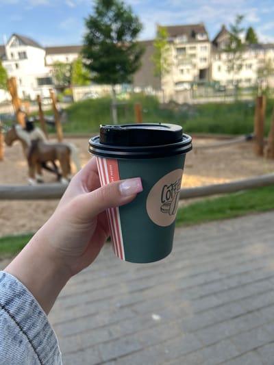 In Waldbröl, Germany, a person's hand holds a dark green disposable coffee cup with a black lid. The cup has a label with red and white stripes and a circular brown sticker featuring a coffee bean illustration and the word "Coffee" in a script font. In the background, a playground with a sand pit and wooden structures is visible, including a toy horse. Residential buildings and trees line the horizon under a partly cloudy sky. The ground in the foreground is paved with grey rectangular stones.