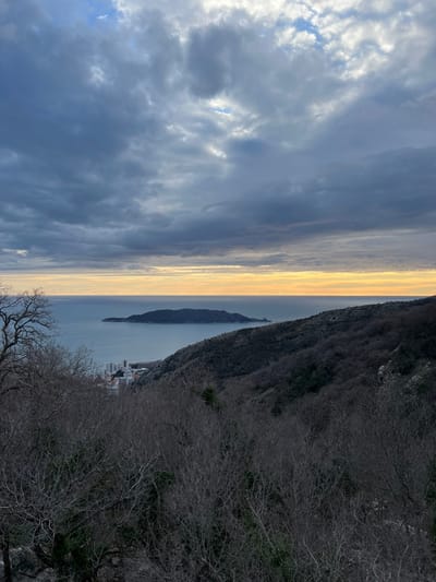 The image displays a panoramic landscape captured from a high vantage point, looking out over a body of water at either sunrise or sunset. The sky, dominating the upper portion of the image, features dramatic cloud formations. Dark grey and bluish clouds cover much of the sky, with patches of brighter white clouds and hints of blue sky visible, particularly towards the upper left. A striking golden-yellow band of light stretches horizontally across the horizon, where the sky meets the distant water, indicating the time of day.
Below this radiant horizon, a calm body of water, likely a sea or strait, extends across the middle ground. The water reflects the muted light of the sky, appearing a silvery-blue with subtle golden tones near the horizon line. In the mid-distance, a long, narrow, and dark landmass or island is silhouetted against the brighter water and sky, stretching horizontally across the view.
The right side of the foreground is occupied by a steep, dark, and densely vegetated hillside or mountain. The trees and bushes on this slope appear bare, suggesting a winter or early spring season, with their intricate, dark branches creating a textured pattern. At the base of this hillside, along the water's edge, a small cluster of buildings or a town can be seen, appearing diminutive from this elevated perspective.
The overall scene conveys a sense of peaceful grandeur, with the interplay of dramatic clouds, golden light, and vast natural elements. There is no visible text or specific activity taking place; the image is a serene landscape view.