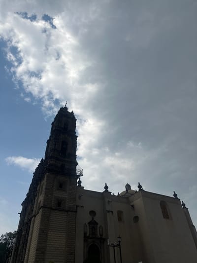 This media file captures a low-angle perspective of a magnificent historical building, likely a church or monastery, set against a dramatic sky. The building, consistent with the architectural style found in colonial Mexico, is ornate and heavily detailed, suggesting Baroque or Churrigueresque influences. A prominent bell tower, intricately carved and dark with age, dominates the central part of the building's visible structure. Decorative finials, round and arched windows, and elaborate stone patterns adorn the facade, which appears to be a light, weathered stone. Portions of a dome are also visible behind the main structure.
The sky occupies a significant portion of the frame. It is a mix of overcast grey clouds, suggesting variable weather, and patches of bright blue sky, particularly towards the bottom and center, indicating breaks in the cloud cover. The lighting is natural, suggesting daytime, possibly late morning or afternoon, with diffused light due to the clouds.
The scene depicts a static view of architecture, with no people or specific events taking place. The grandeur of the building implies a location of historical and cultural significance, aligning with the context of Tepotzotlán, Mexico, renowned for its colonial-era religious structures like the former Colegio de San Francisco Javier (now Museo Nacional del Virreinato). A minimal amount of dark tree foliage is visible at the very bottom right corner. There is no visible text in the image.