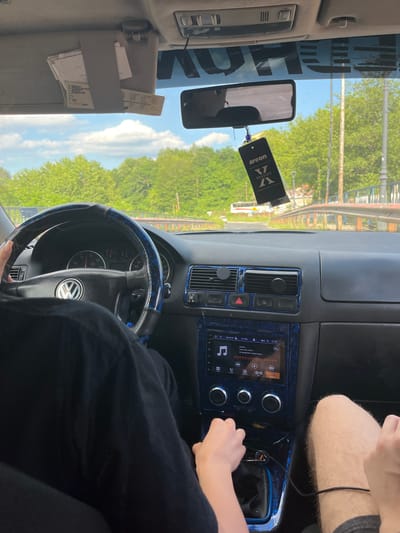 The image shows the interior of a car, likely a Volkswagen Golf, with two individuals inside. The person in the foreground is driving, with their hands on the steering wheel and gear shift. The steering wheel has a custom blue and black cover. The dashboard features an aftermarket infotainment system displaying music details and the time. A car air freshener, labeled "areon X VERSION," hangs from the rearview mirror. The sun visor above the driver is open, revealing several documents. Outside the car, a tree-lined road and a bridge are visible under a cloudy sky. The scene suggests a journey through a rural or suburban area, potentially in the city of Srednogortsi, Bulgaria, given the provided location context.
