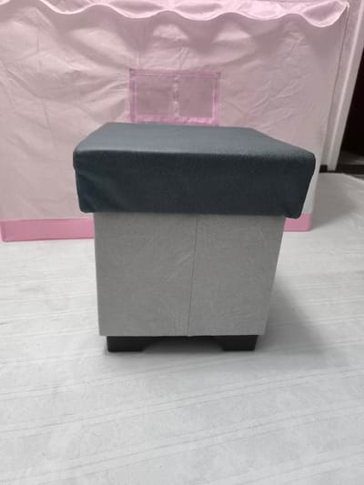This media file showcases a close-up, eye-level view of a square ottoman with a dark gray cushioned top and a light gray textured base. The ottoman has a sturdy appearance, supported by four dark, blocky feet.
In the background, a large, soft pink tent-like structure is visible, featuring a scalloped edge along its top and a transparent rectangular window with a pink trim. The tent is made of a lightweight, possibly fabric material, with visible creases and folds suggesting its pliable nature. A thin, horizontal pink band is present along the lower edge of the tent.
The scene is set on a white, textured surface, possibly a rug or fabric floor covering, with subtle linear patterns. The lighting is even, suggesting an indoor setting with diffused light, and there are no strong shadows. The overall impression is one of a clean, domestic environment. No people are visible in the frame. The image does not contain any visible text. The context provided indicates the location as Kokand, Uzbekistan.