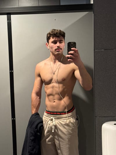 A male subject is depicted from the mid-thighs up, taking a mirror selfie in an indoor environment, likely a gym changing room or public restroom facility in Chantepie, France. The subject has dark, curly hair and a prominent mustache. He is shirtless, displaying a muscular physique, and is wearing light beige sweatpants with an exposed white drawstring. A red and green striped band of underwear is visible above the sweatpants. White wired earbuds are worn, connected to the black smartphone held in his left hand. A silver chain necklace with a small cross pendant hangs around his neck. His right arm is holding a dark blue or black garment, possibly a jacket or hoodie. The smartphone has a textured, black case with a swirling pattern. The background consists primarily of a light gray wall reflected in the mirror, with a darker gray textured panel visible on the right side. A black vertical frame or partition is present on the left. On the far right, a portion of a white wall-mounted fixture, resembling a urinal or toilet cistern, is partially visible. The lighting is bright and even.