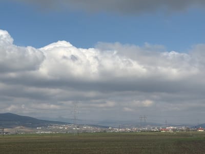 A photograph taken in Alba Iulia, Romania, depicts a landscape under a cloudy sky. The sky is segmented into areas of clear blue and large formations of gray and white cumulus clouds, creating a dramatic contrast. In the foreground, a brown agricultural field stretches toward the middle ground. The field exhibits regular patterns indicative of tilled soil or planted rows.

The middle ground is characterized by the presence of a town or city, indicated by the cluster of buildings and structures. The built environment appears to blend into the natural surroundings. Power lines supported by tall metal pylons traverse the landscape, cutting across the open fields and the skyline. The pylons are spaced at intervals and diminish in size with distance, conveying perspective.

The background features a low mountain range silhouetted against the sky, adding depth to the composition. The general light conditions suggest overcast weather, casting a subdued tone over the entire scene. There are no people visible in the image.