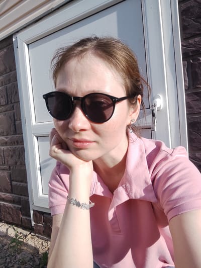 A woman in sunglasses and a pink polo shirt poses for a selfie outdoors. She is resting her head on her hand, which features a silver bracelet. Behind her is a white door with keys hanging from the handle and a wall with brown brickwork. The location is Malaya Purga, Russia.