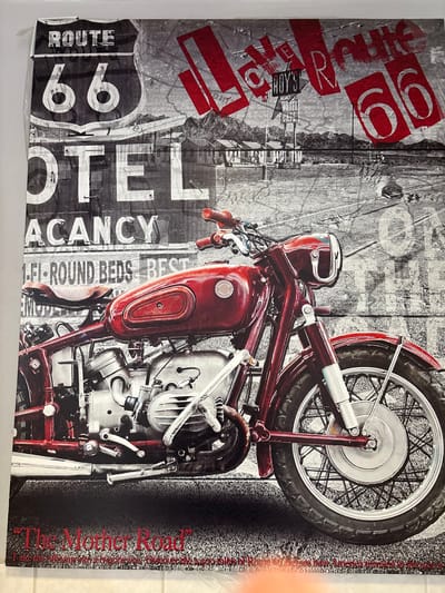 This image displays a vertically oriented rectangular artwork, presumably a print or canvas, present in West Felton, United Kingdom. The artwork's primary subject is a vibrant deep red vintage motorcycle, prominently featured in the foreground, angled to the right. Details include its chrome engine components, exhaust pipes, a textured brown leather seat, and spoked wheels with a light-colored hub on the front.

The background of the artwork is rendered in grayscale, depicting a classic American Route 66 roadside environment. In the upper left quadrant, a large "ROUTE 66" shield sign is visible, along with a "MOTEL VACANCY" sign directly beneath it. Further text on signs includes "1-FI-ROUND BEDS" and "BEST". In the upper right section, distressed red text reads "Love ROUTE 66", accompanied by a sign for "ROY'S VACANCY MOTEL CAFE". A low-rise motel complex with multiple units extends horizontally in the mid-ground, set against a backdrop of distant mountains under a cloudy sky. The bottom of the artwork features the red text "The Mother Road". No individuals are present in the depicted scene within the artwork.