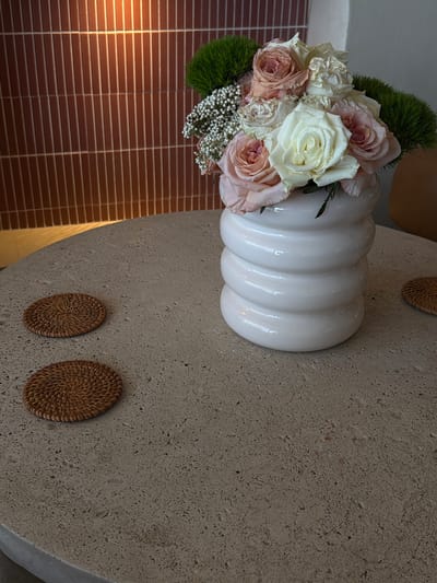 A close-up, eye-level shot shows a bouquet of roses in various shades of pink and white, nestled in a glossy white vase with a ribbed design. The vase sits on a textured, light-colored round table. Two round, woven coasters are placed on the table to the left of the vase. Behind the table, a wall with vertical, reddish-brown tiles is visible, lit from the top. The overall setting suggests an indoor environment, possibly a restaurant or cafe, in Fort Lauderdale, Florida, United States of America. No people are present in the image.