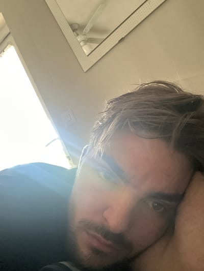 This close-up, low-angle selfie captures a man indoors, likely in Rafz, Switzerland. His face, angled upwards and slightly to the left, dominates the right and central portions of the frame. He has light brown or blonde, somewhat messy hair, and his right eye is clearly visible, looking directly at the camera. A subtle beard or stubble is present around his chin and upper lip. He appears to be wearing dark clothing on his upper body.

The background shows a light-colored ceiling. To the far left, a mirror with a textured, decorative frame reflects a ceiling fan or light fixture. Another rectangular fixture or vent is visible on the ceiling towards the center. A very bright light source, possibly a window, creates significant glare across the lower-right and central-bottom parts of the image, illuminating parts of the man's hair and obscuring some details. The overall lighting is strong and high-contrast. The man's expression is neutral and direct. No text is visible in the image.