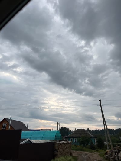 The image shows a rural scene in the city of Zuevo, Russia, under a cloudy sky. In the foreground, there are dark fences and a dirt path. To the left, a brown wooden house with a grey roof and a chimney is visible. Behind it, a long, curved structure with a teal-colored translucent roof is prominent. Further to the right, a smaller, older house with a grey tiled roof and light blue siding is situated amongst trees. A utility pole with power lines runs diagonally across the mid-ground. The background consists of a dense line of dark green trees. The overall impression is one of overcast weather.