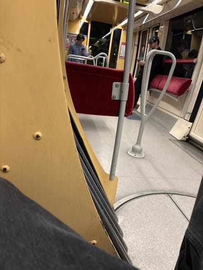 The image captures the interior of a public transport vehicle, likely a tram or an articulated bus, at a low-angle, first-person perspective. The setting appears to be during the evening or night, with artificial lighting illuminating the cabin.
Two individuals are visible. In the mid-ground, a person wearing a dark cap and jacket stands, looking down at a mobile phone. A reflection of this person is seen in the window behind them. Closer to the foreground on the left, another person is seated, wearing a light-colored shirt and headphones, also engrossed in their mobile phone. Red upholstered seats are scattered throughout the vehicle, with one prominently featured in the mid-ground.
The vehicle's interior features light grey flooring and creamy yellow/beige walls and structural elements. Numerous grey metal grab handles and poles are present; a distinctive white U-shaped handle is in the mid-ground, and a horizontal bar crosses the foreground. A circular metallic track on the right side of the floor suggests an articulation joint, characteristic of trams or long buses. Part of the viewer's leg, clad in dark denim, is visible in the lower right corner.
A sign on the upper left displays partially visible text: "B Hoer", serving as a location cue consistent with Hoenheim, France. The scene depicts a common commuting or travel scenario within public transport.
