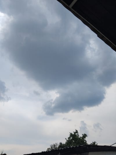 The image shows an overcast sky with large, dark clouds dominating the upper left portion. Towards the right, the sky is lighter, with some scattered white clouds. A dark, textured roofline extends from the top left corner down into the frame, casting a shadow or being a part of a structure. On the far right, a sliver of a dark roofline is visible, with green foliage from a tree peeking out from behind it. The overall scene suggests an outdoor setting, possibly a residential area given the presence of roofs and a tree. The lighting indicates it is daytime, likely afternoon or late morning, with the weather appearing to be cloudy or on the verge of rain. There are no people or specific activities depicted. No text is visible in the image.