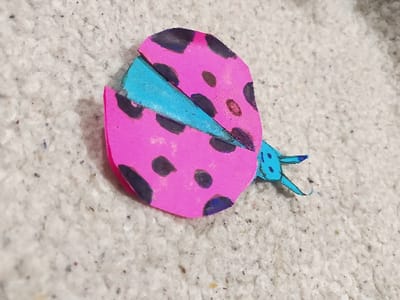 The scene, located in Harasta, Syria, depicts a single, stylized paper craft item resembling a ladybug, positioned diagonally on a light-colored, textured surface. The craft features an oval-shaped body, predominantly pink, adorned with multiple irregular black spots. A central segment, likely representing underwings or a thorax, and a small head with two antennae are colored light blue. The craft exhibits visible folds, cuts, and edges consistent with paper construction. The background is a coarse, off-white or light beige fibrous surface, consistent with a carpet or rug, displaying scattered dark particulate matter and individual fibers. The lighting is soft and even, illuminating the subject and its immediate environment.