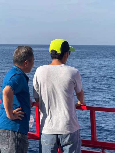 Two individuals stand on the deck of a vessel, facing away from the camera and towards the open sea. The person on the left, wearing a blue polo shirt and dark jeans, has their left hand on their hip. The person on the right, wearing a grey t-shirt and blue jeans, has a bright yellow and black baseball cap on and is leaning on a red railing. The sea is a deep blue with visible ripples, and the sky is a pale, cloudless blue. The location is identified as the city of Dong Son, Vietnam.