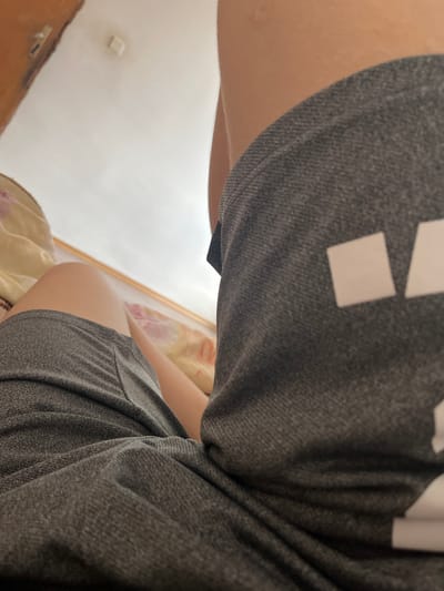 The image captures an indoor scene from a low-angle perspective, looking upwards. The primary subject is a person, partially visible through their legs and thighs, suggesting they are lying down or seated on a bed. They are wearing casual gray clothing, possibly shorts or sweatpants, made of a textured fabric. On the upper part of the gray fabric, a clear white square shape is visible, along with parts of another larger white shape, which could be elements of a logo or pattern.
In the background, a light-colored wall or ceiling is prominent, with what appears to be a small, square, recessed light fixture or similar object embedded in it. A wooden doorframe or furniture is partially visible on the left side of the frame. Below the person, a patterned bedding or mattress is seen, featuring light yellow and pink/purple floral or abstract designs.
The scene conveys a relaxed atmosphere, likely taking place during the daytime given the soft, ambient light illuminating the room, possibly through a window not directly visible. The location context for this interior scene is Gorno Dryanovo, Bulgaria. No discernible text is visible in the image.
