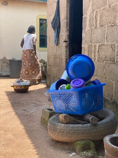 In a courtyard in the city of Daniel, Nigeria, a woman with braided hair stands with her back to the camera, wearing a white t-shirt and a patterned skirt. She is near a beige building with a green-framed window. In the foreground, a collection of blue plastic containers are stacked. A blue basket holds several items, including purple and green lids and a large blue bowl rests on top of it. This basket and bowl are placed on a worn tire, which is itself situated on a rough, sandy ground. Other objects visible include a dark grey bin and a pot filled with plastic bottles and other items. The scene suggests a domestic setting with items arranged for storage or use.