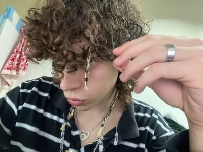A close-up, tilted, portrait-style photograph depicts a person with curly brown hair. The person is wearing a black and white striped shirt and several necklaces, including one with a silver heart-shaped charm. The person's right hand is visible, with a silver ring on the ring finger. Two strands of hair are braided with what appears to be a light-colored material, possibly yarn or string, held in place with black bands. The background includes a glimpse of a curtain or fabric with a pink and red pattern. There is no discernible information about the location being Bahía Blanca, Argentina.
