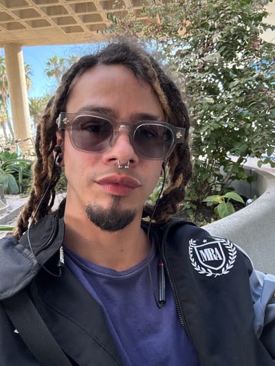 A man with dreadlocks and sunglasses is outdoors, possibly in Montaña Roja, Spain. He has a nose ring and a short beard. He is wearing a blue shirt and a black jacket with a white emblem on the shoulder. He also has wired earbuds connected to his phone. The background includes greenery, architectural features, and glimpses of the sky.