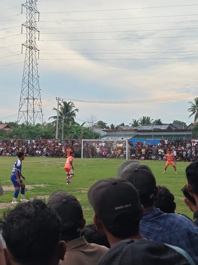 This image depicts a soccer match in Desa Meunasah Blang, Indonesia. A large crowd is gathered to watch the game, many seated and standing along the sidelines. Two players in orange uniforms are visible on the field, one in the foreground and another near the goal. A player in a blue and white uniform is also present. The game is being played on a grassy field with a soccer goal and net. In the background, there are trees, buildings, and a tall electricity pylon. The sky is overcast with some clouds. The perspective of the image is from the crowd, looking towards the field.