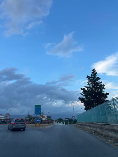 The image depicts a view from a vehicle driving on a paved road in Manfredonia, Italy. In the foreground, a dark gray station wagon with the license plate "FW 954LZ" is visible. The road appears to be an intersection with multiple road signs. One sign indicates "autostrade" with an arrow pointing upwards, while others point towards "S. Giovanni Rotondo 30," "M.te S. Angelo 33," "FOGGIA 36," and "VIESTE 68." There is also a sign for "MANFREDONIA 4." To the right of the road, a tall, dark green coniferous tree stands against a partly cloudy blue sky. A light turquoise metal fence runs along the right side of the road. In the background, buildings and hills are visible. The overall scene suggests a cloudy day with twilight approaching.