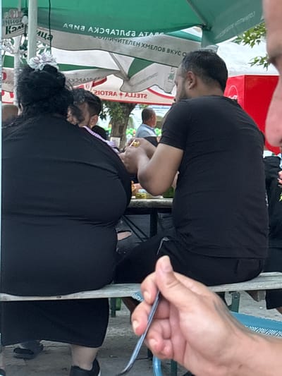 The image depicts an outdoor dining scene in Kostievo, Bulgaria, featuring several individuals seated at tables under large canopies during daylight hours. In the mid-ground, viewed from behind, are two individuals seated on a bench at a table.
On the left, a person, likely an adult female, is wearing a black, form-fitting garment and has dark hair styled with a light-colored, possibly white or pale pink, floral accessory on the back of their head. Their posture indicates they are seated facing away from the camera.
To their right, an adult male is also seated, wearing a black short-sleeved t-shirt and dark trousers. He has short, dark hair with visible grey streaks on the sides. His hands are engaged over the table, with one hand holding a small, reflective, possibly metallic object like a ring, while the other hand is positioned near it. A green bottle is partially visible on the table between the two individuals.
In the extreme foreground, a light-skinned hand, likely belonging to the photographer, is visible holding the handle of a silver utensil (possibly a fork or spoon).
The background features several large outdoor umbrellas providing shade. One prominent umbrella displays Cyrillic text, which, when unreversed, reads "АРНА АЖЯЧАТПЯА А" and below it "ТОЛИЧИТЕЛЕН ХМЕЛОВ ВКУС О", indicating a "delicious hops taste". Another umbrella shows the reversed text "2IОTЯA+A773T2", which unreversed reads "STELLA+ARTOIS", identifying a beer brand. Further in the background, other patrons are visible, including a man in a light blue shirt on the far left and another man in a light shirt under an umbrella in the center-background. A red box-like structure, possibly a cooler or vending unit, with a white logo is visible on the far right. The ground is paved, and trees and foliage are visible in the distance, indicating an outdoor, possibly park-like or street-side, setting.