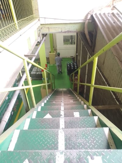 An indoor industrial staircase is depicted in a downward view, with a person walking away from the camera at the top of the stairs. The staircase is made of metal with green diamond plating on the steps and white painted risers. A yellow metal handrail runs along the right side of the stairs. The environment appears to be a factory or warehouse with green painted floors, yellow support beams, and corrugated metal walls. In the background, there are stacks of black plastic bottles and some crates. The location is identified as Sukaresmi, Indonesia.