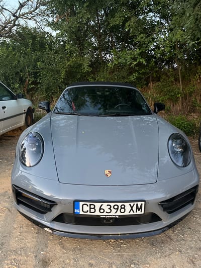 The media file displays a close-up, top-down view of the front of a grey Porsche. The car is parked on a dirt and gravel surface, with lush green foliage and trees framing the left side of the image. The car's headlights are illuminated, and its front bumper features black accents and air vents. A license plate with the inscription "CB 6398 XM" and the Bulgarian flag is prominently displayed. Below the license plate, a website address "www.porsche.bg" is visible. The scene appears to be outdoors, likely in a rural or natural setting, given the presence of trees and the unpaved ground. The lighting suggests it could be late afternoon or early evening, with warm tones reflecting on the car's surface. No people are visible in the image. The overall impression is one of a parked luxury vehicle in a natural environment.