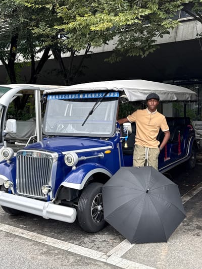 The image captures an outdoor scene in the Olympic Sailing Marine Culture Tourist Area, Shinan District, Qingdao, China. A male subject, appearing to be in his twenties, with dark skin, stands next to a dark blue, vintage-style electric tourist vehicle. He wears a beige polo shirt with a darker collar, light olive green cargo shorts with white drawstrings, and a dark flat cap; a watch is visible on his left wrist. His right elbow rests on the vehicle's side, and his left hand is in his pocket.
The dark blue vehicle features a chrome grille, bumper, and circular headlight surrounds. A light-colored canvas canopy covers its passenger area. Above the windshield, a blue banner displays Chinese characters, "奥帆海洋文化旅游区" (Olympic Sailing Marine Culture Tourist Area). A small bell is affixed to the right side mirror support. The vehicle's tires have black rims with silver accents.
A black, fully open umbrella lies on the asphalt pavement in front of the blue vehicle's right wheel. To the left and slightly behind the blue vehicle, a portion of a white electric shuttle is visible. The background consists of trees with green foliage and a modern building with a dark facade. The ground is asphalt with faded white parking lines. The lighting suggests an overcast or shaded daylight environment.